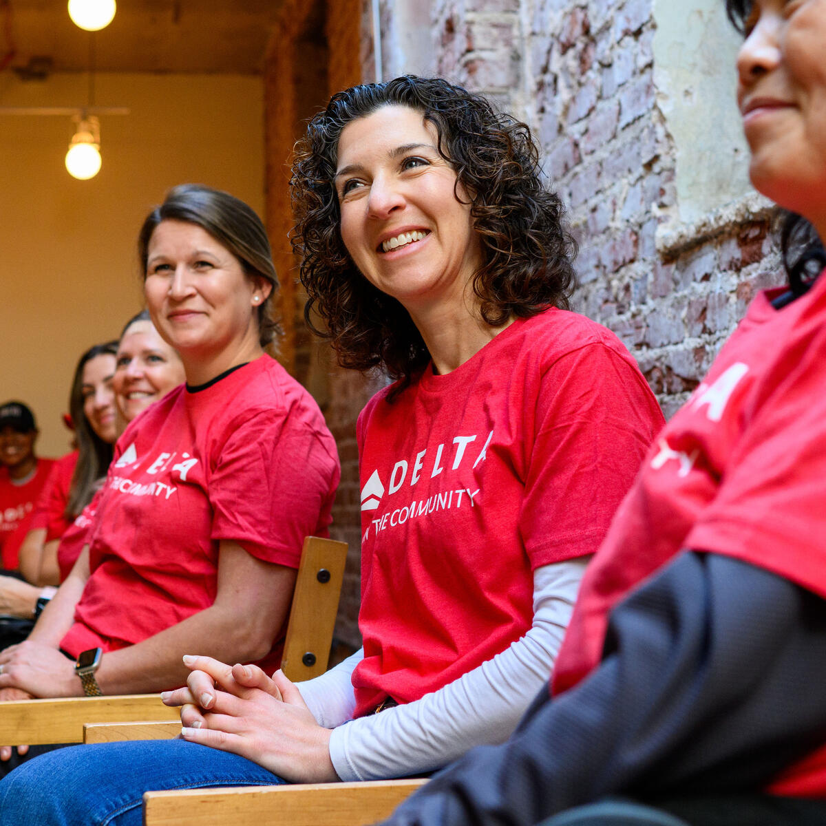 Delta volunteers wearing red “Delta in the Community” shirts sit side by side on chairs inside an indoor space with exposed brick walls during a community event.