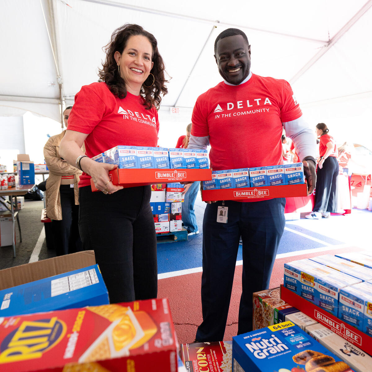 Two Delta volunteers wearing red “Delta in the Community” shirts carry boxes of snack food at a community packing event inside a large tent.