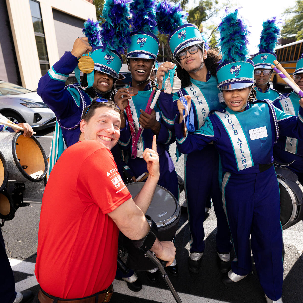 Delta volunteer in a red shirt interacts with a marching band in teal and purple uniforms, with drums and feathered hats, during an outdoor community event.