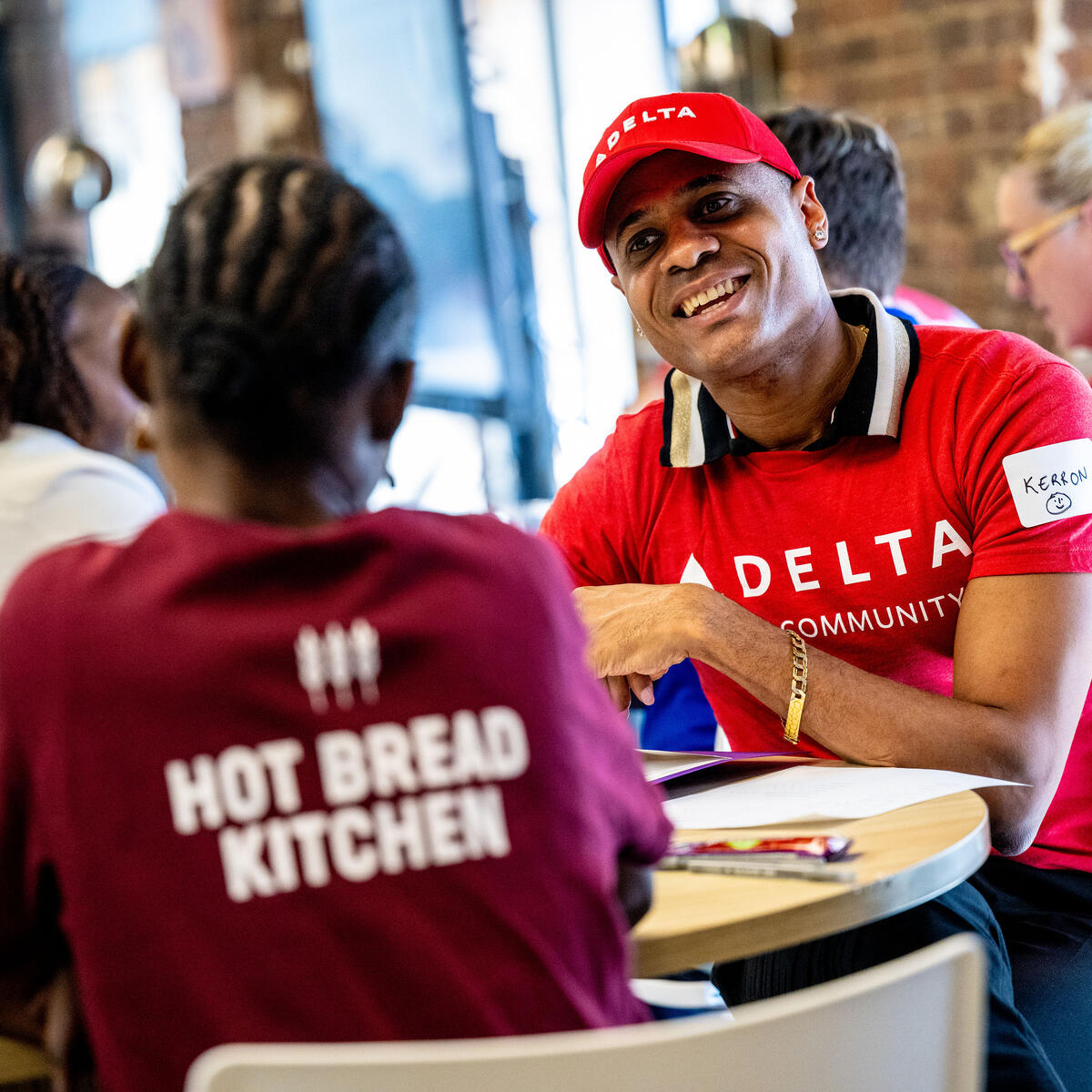 Delta volunteer in a red “Delta in the Community” shirt sits at a table speaking with a community member wearing a “Hot Bread Kitchen” shirt inside a bright indoor space.