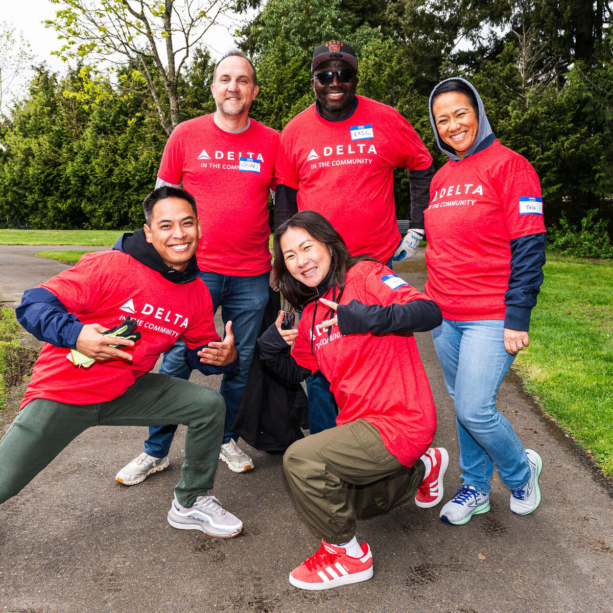A group of Delta volunteers wearing red “Delta in the Community” shirts pose together on a park path, surrounded by green grass and trees.