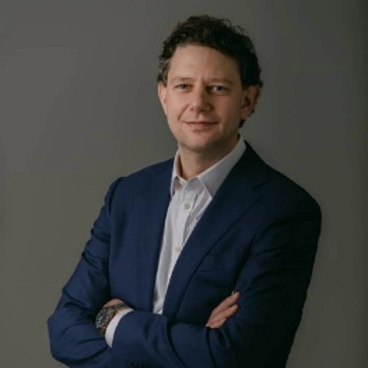 Studio portrait of a person in a dark suit and white shirt, standing with arms crossed against a plain gray background.