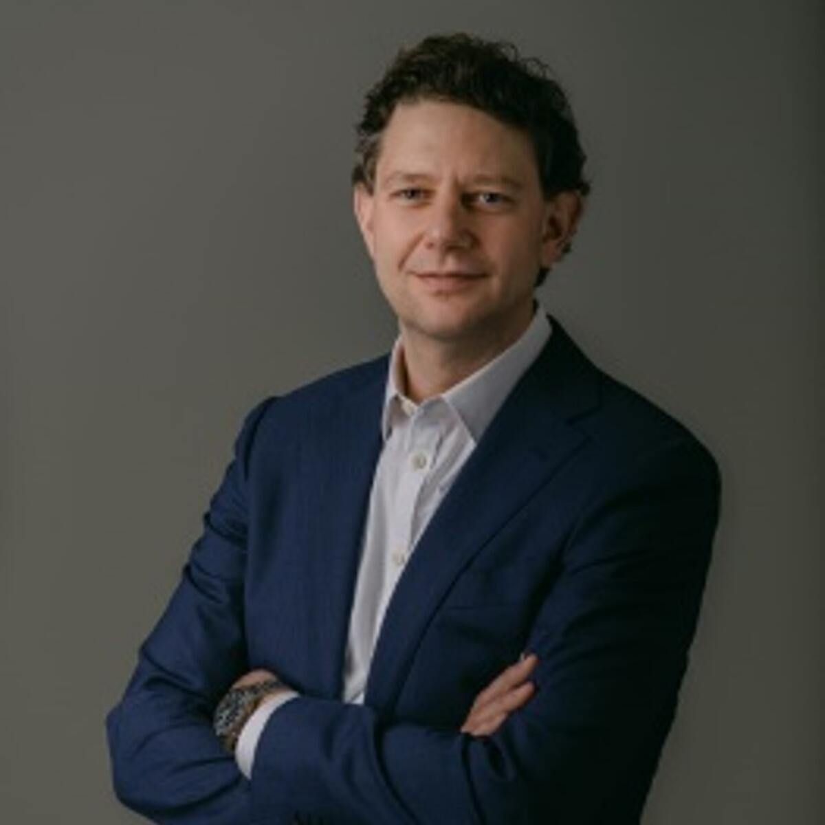 Studio portrait of a person in a dark suit and white shirt, standing with arms crossed against a plain gray background.