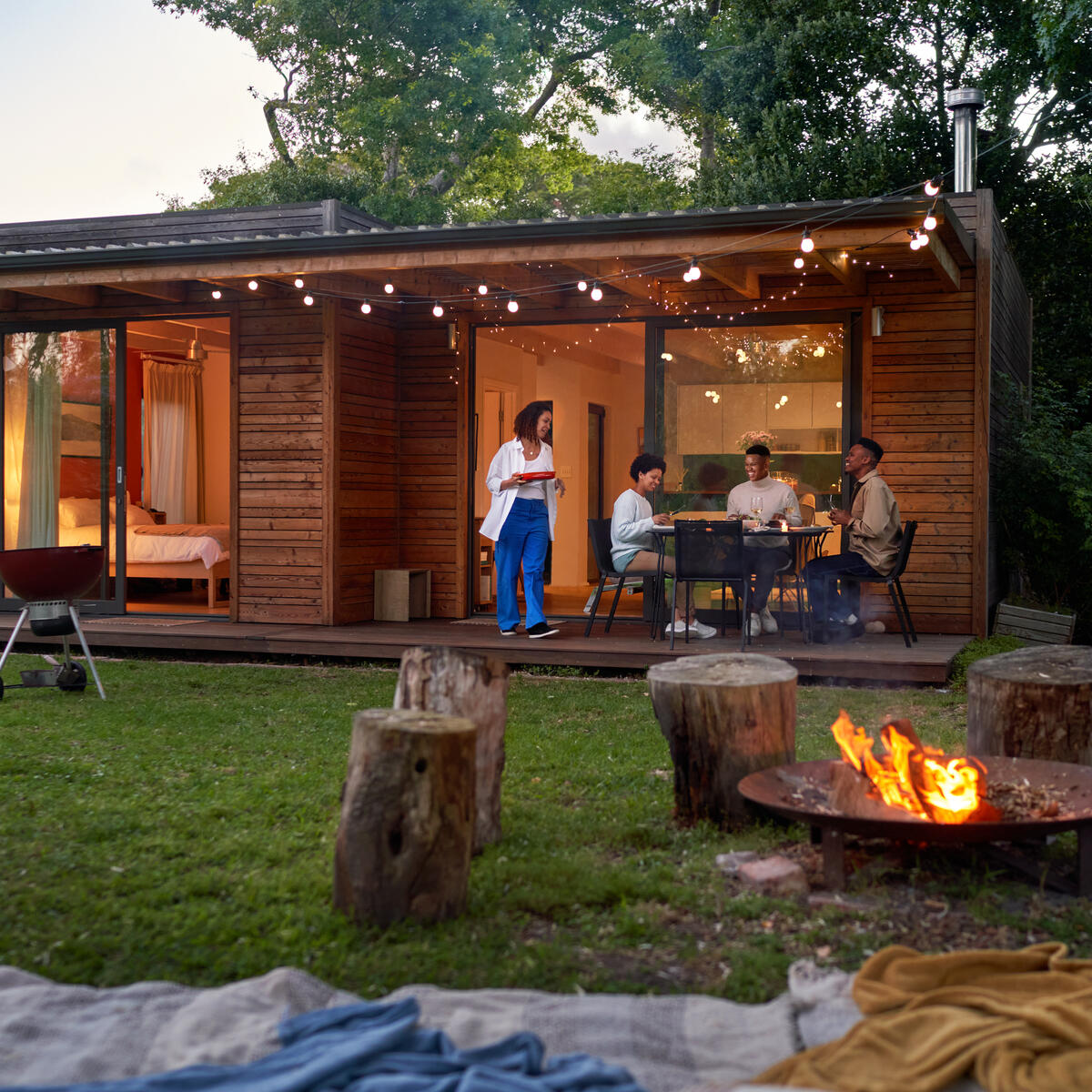 People dining outside a modern wooden cabin at dusk, with string lights, a fire pit and trees surrounding the yard.