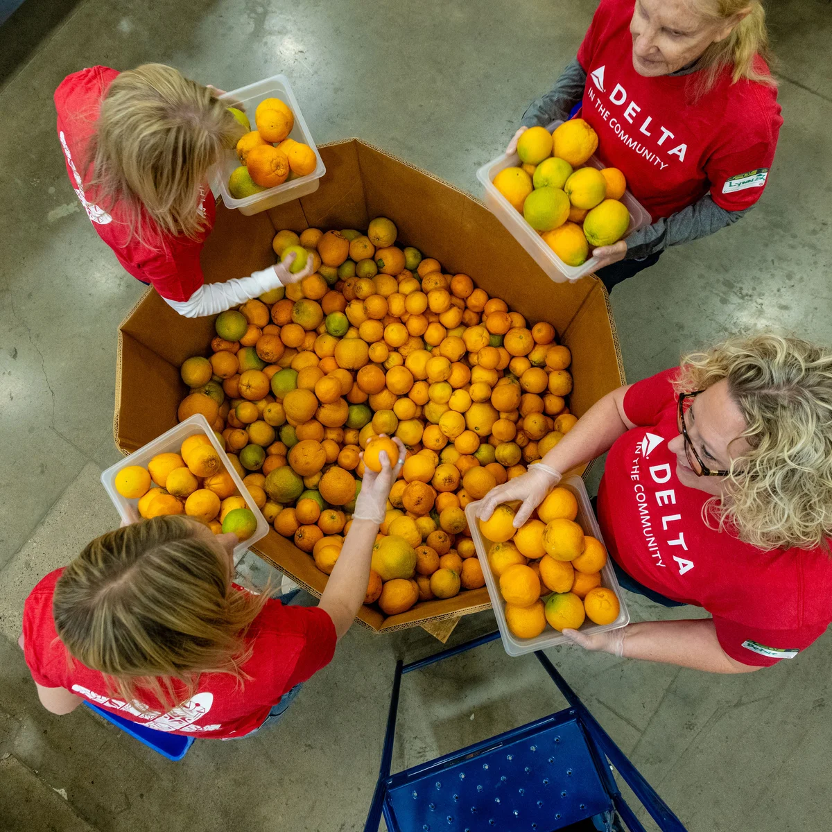 Overhead view of Delta volunteers wearing red “Delta in the Community” shirts sorting oranges into containers around a large bin inside a warehouse setting.