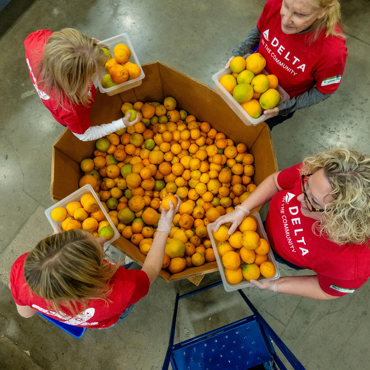 Overhead view of Delta volunteers wearing red “Delta in the Community” shirts sorting oranges into containers around a large bin inside a warehouse setting.
