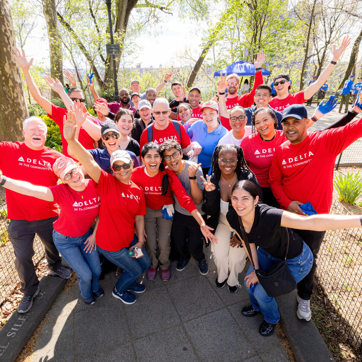 A large group of Delta volunteers wearing red “Delta in the Community” shirts stand together in a park with arms raised, gathered along a paved path surrounded by trees.
