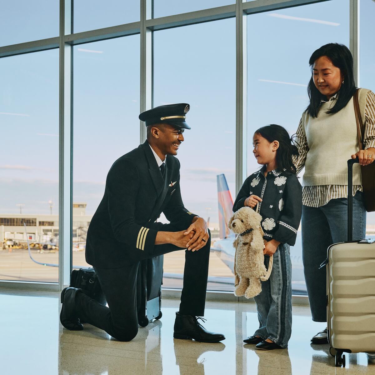 Delta pilot kneeling beside a young child holding a stuffed animal at an airport gate, while an adult stands nearby with rolling luggage; airplane visible through large windows in the background.
