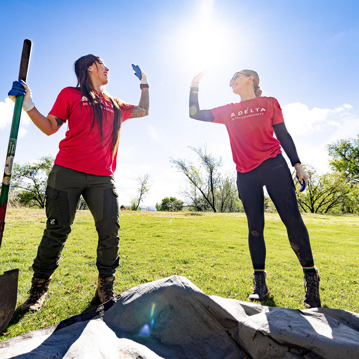 Two Delta volunteers wearing red shirts stand outdoors in a grassy area, raising gloved hands in a high‑five beside a shovel and newly planted tree under a bright sky.