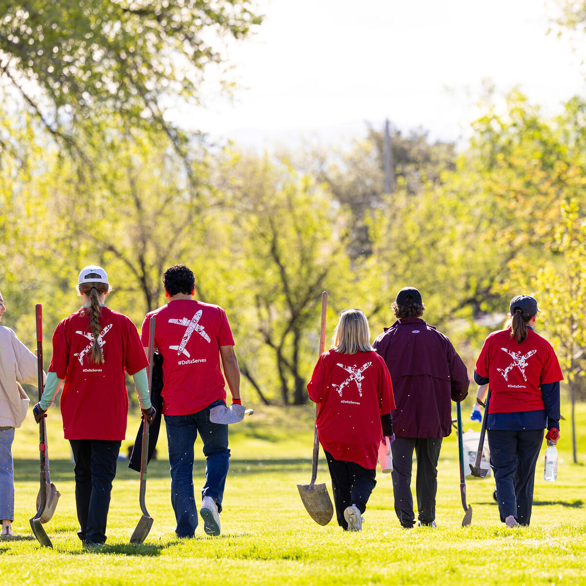A group of Delta volunteers wearing red shirts walk across a grassy park carrying shovels, heading to a tree‑planting activity on a sunny day.