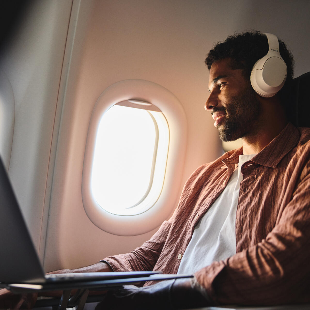 Delta customer wearing headphones works on a laptop while seated by an airplane window, with soft daylight illuminating the aircraft cabin.