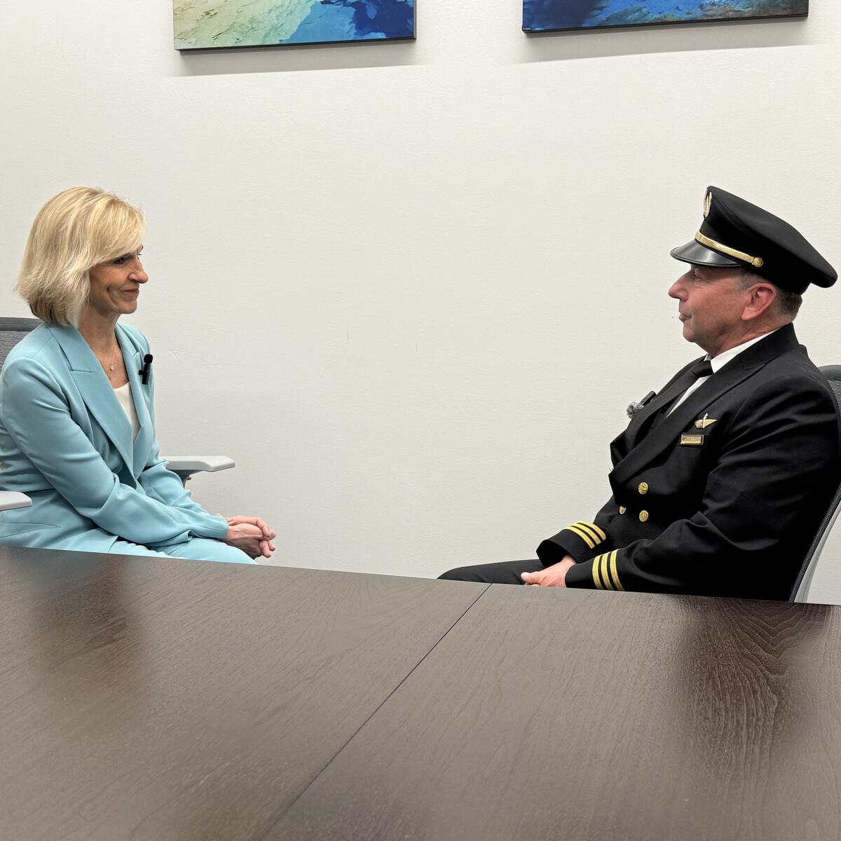 Allison Ausband and Tom Cohan sit facing each other at a conference table, Allison in a business suit and Tom in a pilot uniform.
