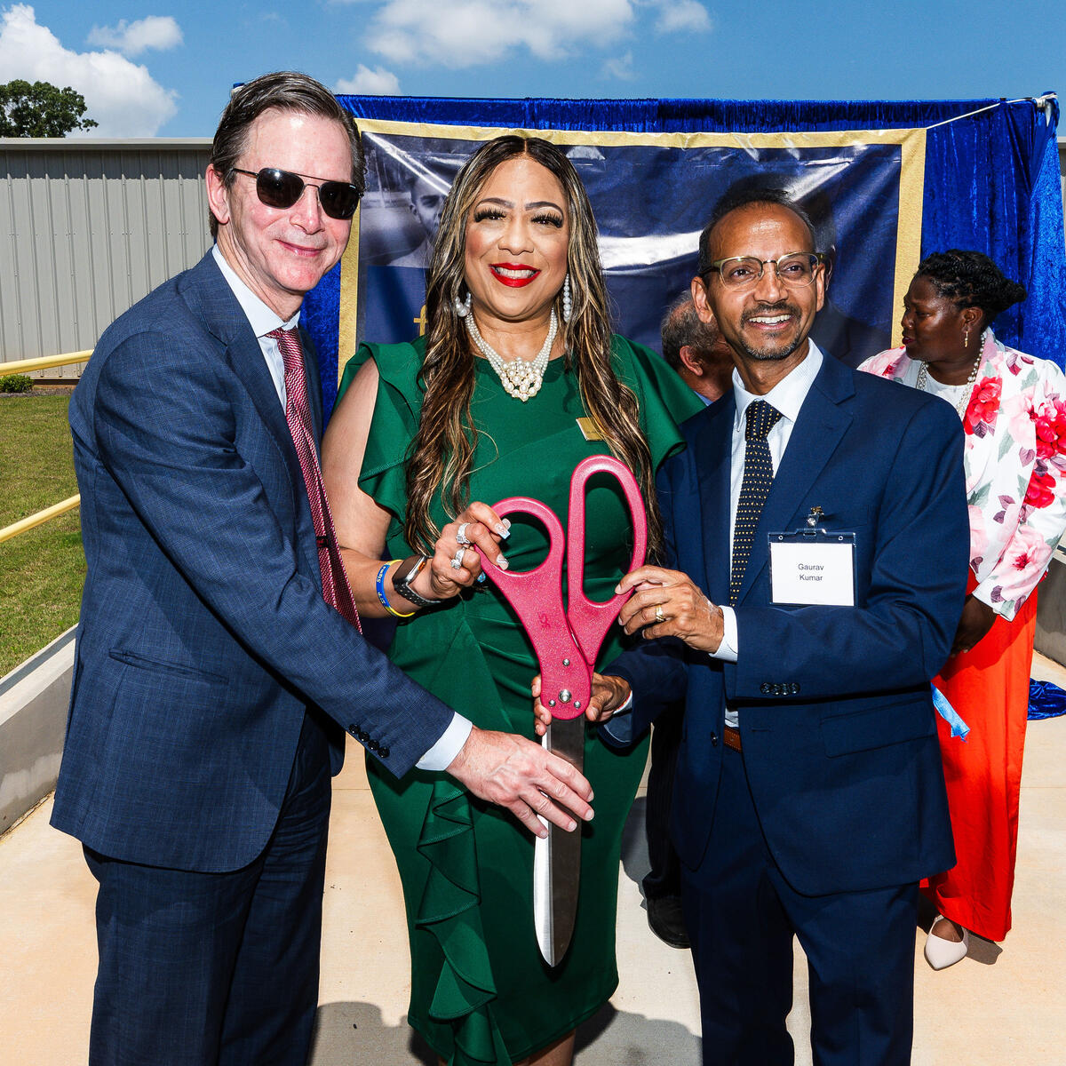 Group of individuals holding oversized ceremonial scissors at an outdoor ribbon-cutting event.
