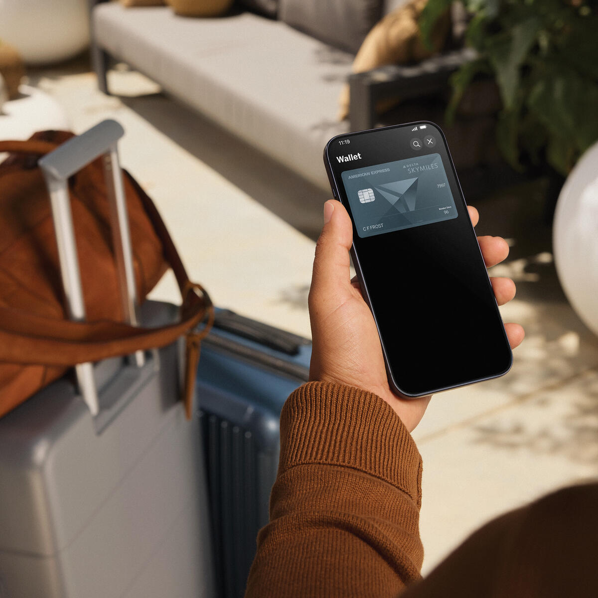 Person holding a smartphone displaying a Delta SkyMiles American Express Card in a digital wallet, with a suitcase and travel bag nearby in a lounge setting.