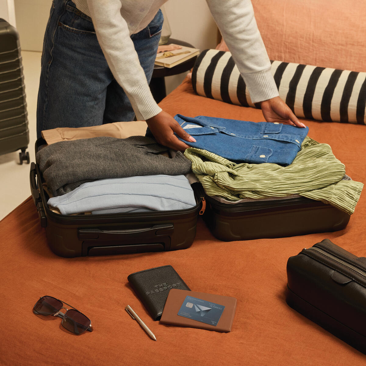Person packing clothes into an open suitcase on a bed, with a Delta SkyMiles American Express Card, wallet, passport, and sunglasses placed nearby.