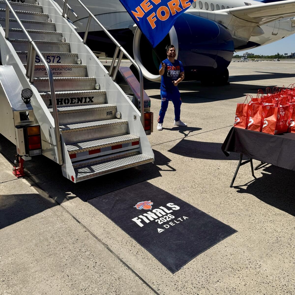 Orange New York Knicks foam finger reading “#1 FAN” sits atop Delta-branded blue towels at an airport display, with travelers and check-in counters in the background.