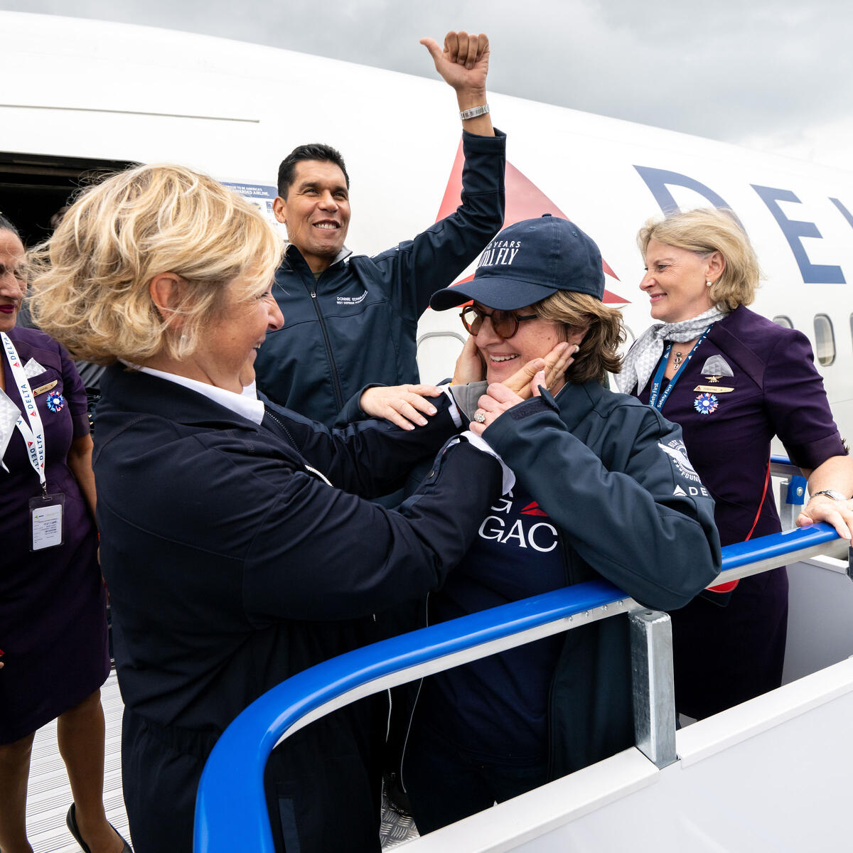 Delta employees and volunteers stand on an airport boarding ramp beside a Delta aircraft, welcoming a passenger as they exit the plane.