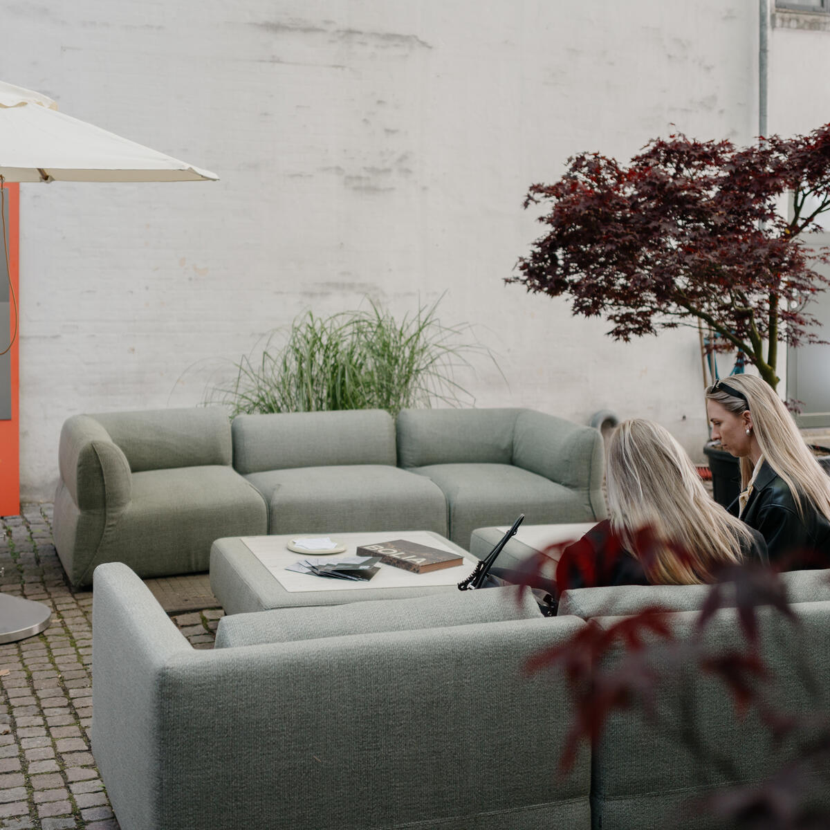 Outdoor courtyard seating area with modular sofas and a low table under an umbrella, where two people sit and talk beside plants and an event information sign.