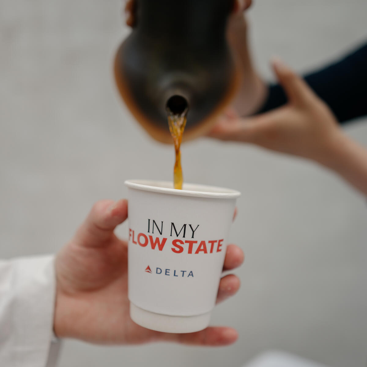 Coffee being poured from a dark ceramic pitcher into a white paper cup printed with “In My Flow State” and the Delta logo.