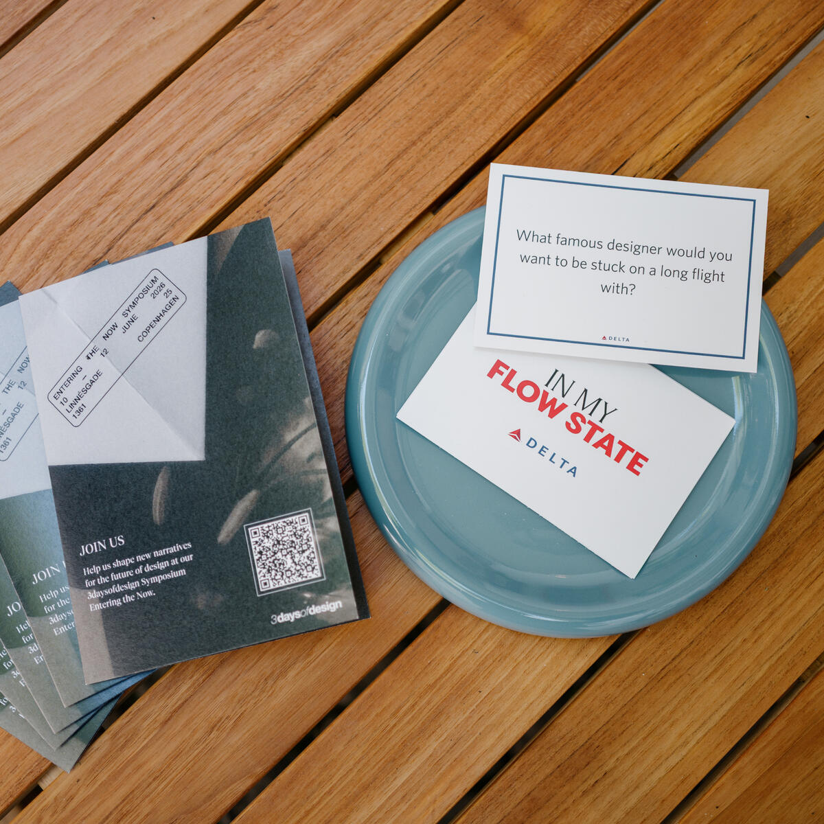 Printed prompt cards and brochures arranged on a wooden table, including a card reading “What famous designer would you want to be stuck on a long flight with?” beside an “In My Flow State” Delta card.