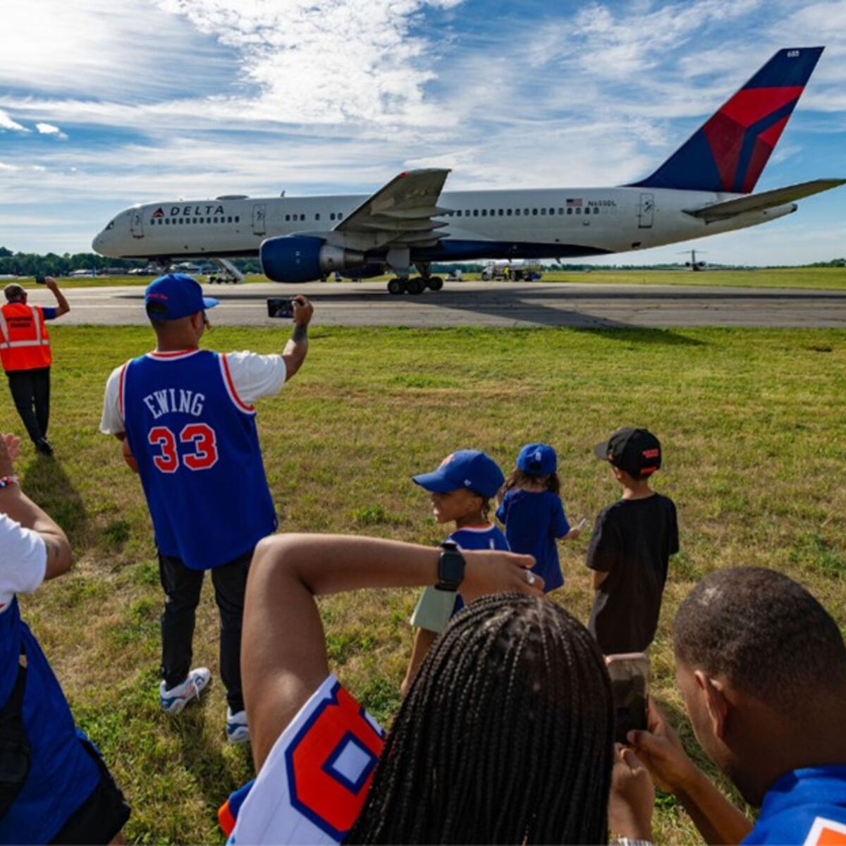 Group of people in sports jerseys standing on grass watching and taking photos of a Delta airplane taxiing on a runway while staff in safety vests stand nearby.