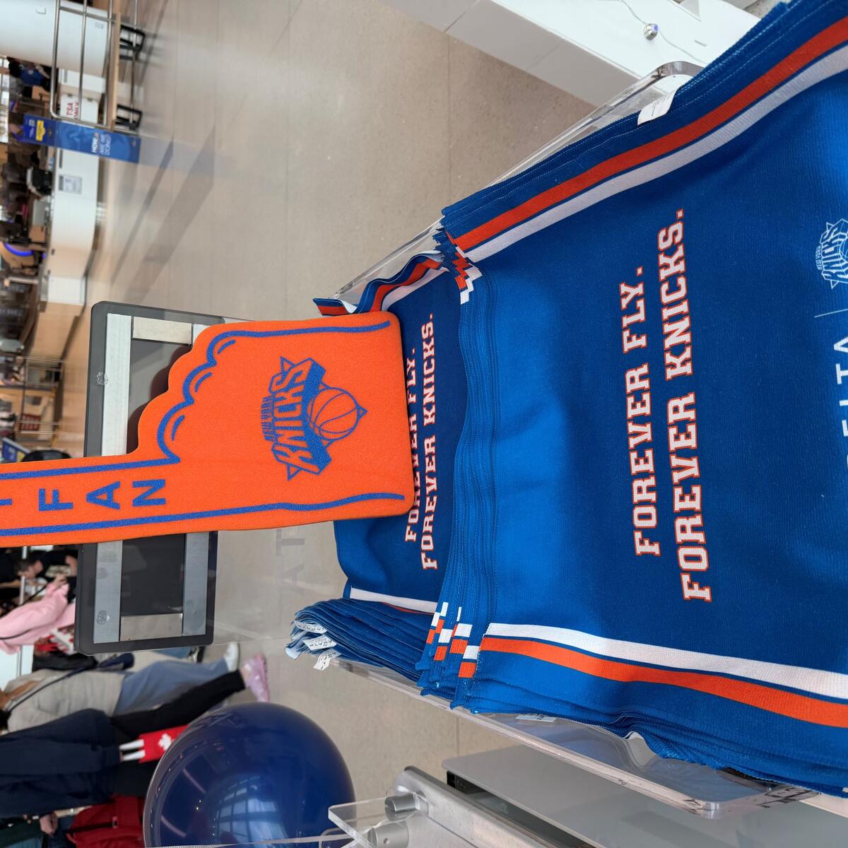 Bright orange New York Knicks foam finger displayed on a stand of blue Delta‑branded rally towels reading “Forever Fly. Forever Knicks” in an airport terminal, with travelers and security area in the background.