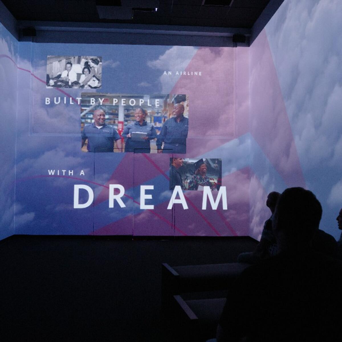 Silhouetted viewers watch an immersive projection of clouds with photos of people and the text “BUILT BY PEOPLE WITH A DREAM.”