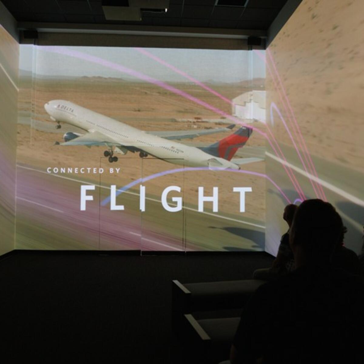 Silhouetted audience in a dark room viewing a multi-wall projection of a plane taking off, with the text “CONNECTED BY FLIGHT” overlaid.