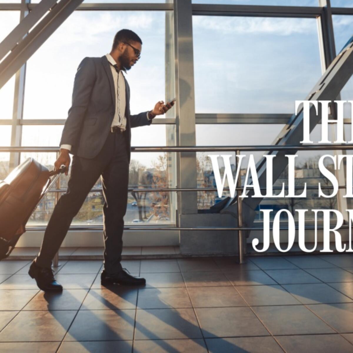 Traveler in a suit walks through a sunlit airport terminal, pulling a rolling suitcase and looking at a phone, with “The Wall Street Journal” text overlaid.