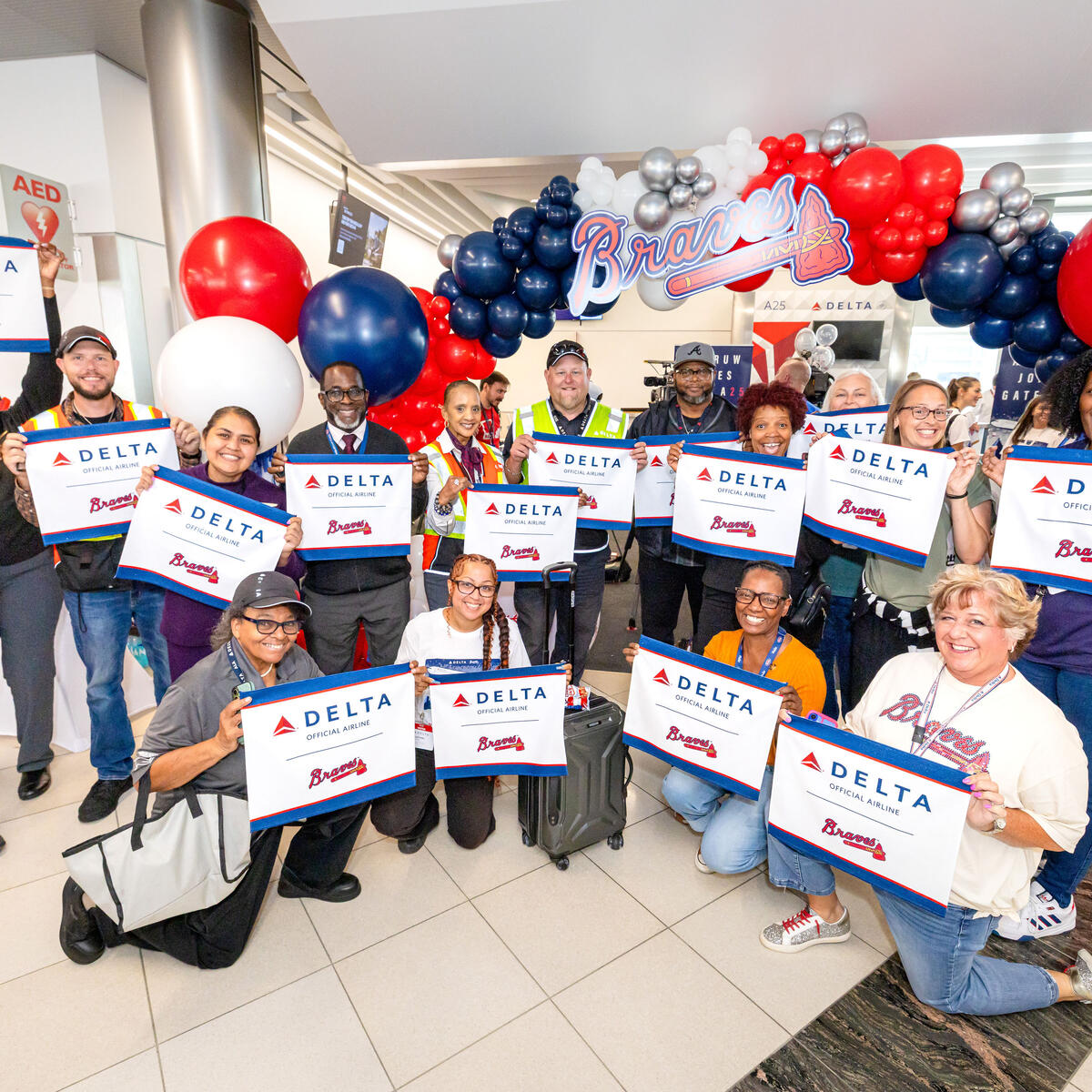 Group of Delta employees and guests pose beneath a Braves-themed balloon arch, holding commemorative Delta and Braves signs during the Gate A25 celebration.