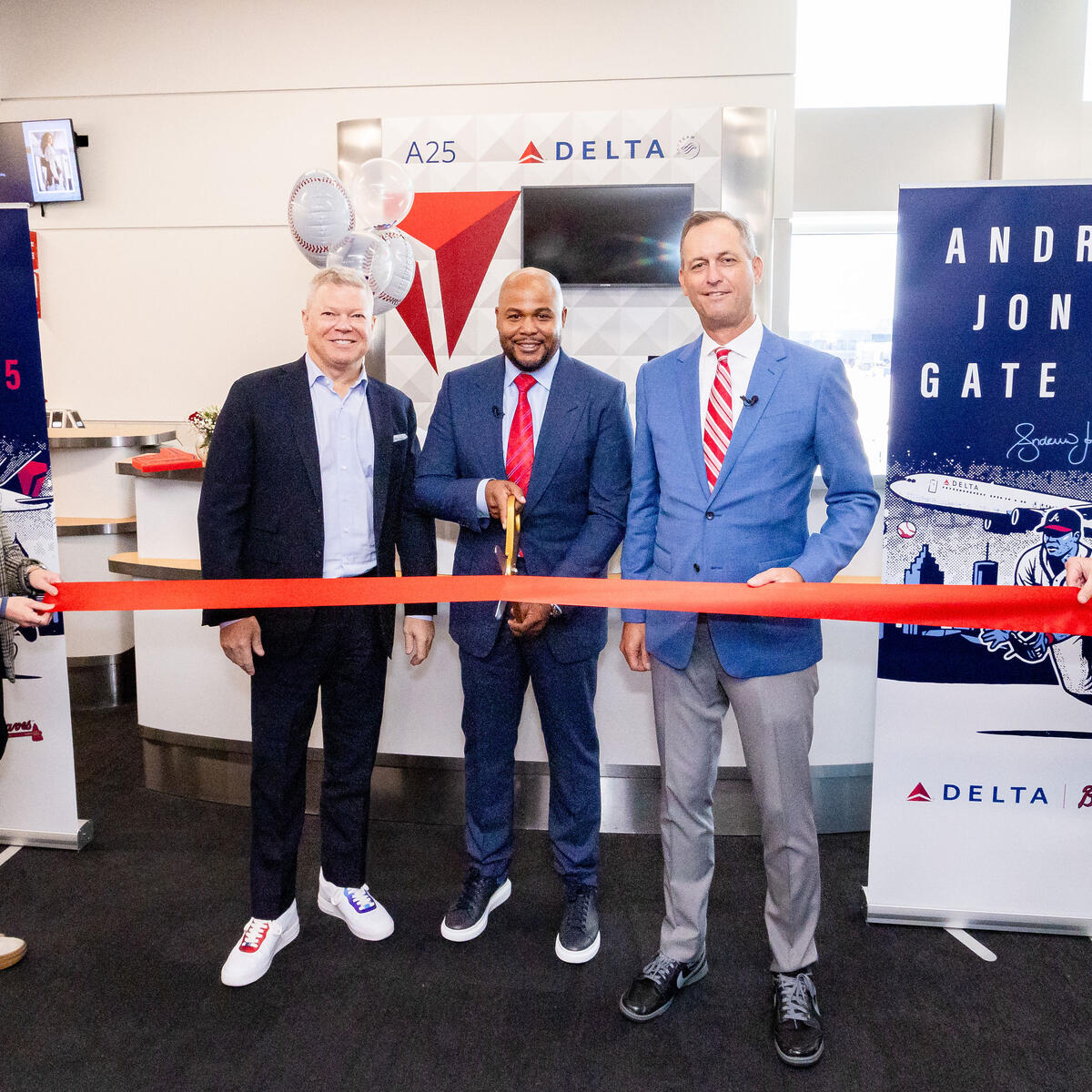 Five people pose at a Delta airport gate ceremony, with the center person holding large scissors to cut a red ribbon. Banners on both sides read “Andruw Jones Gate A25,” featuring baseball-themed artwork and Delta branding.