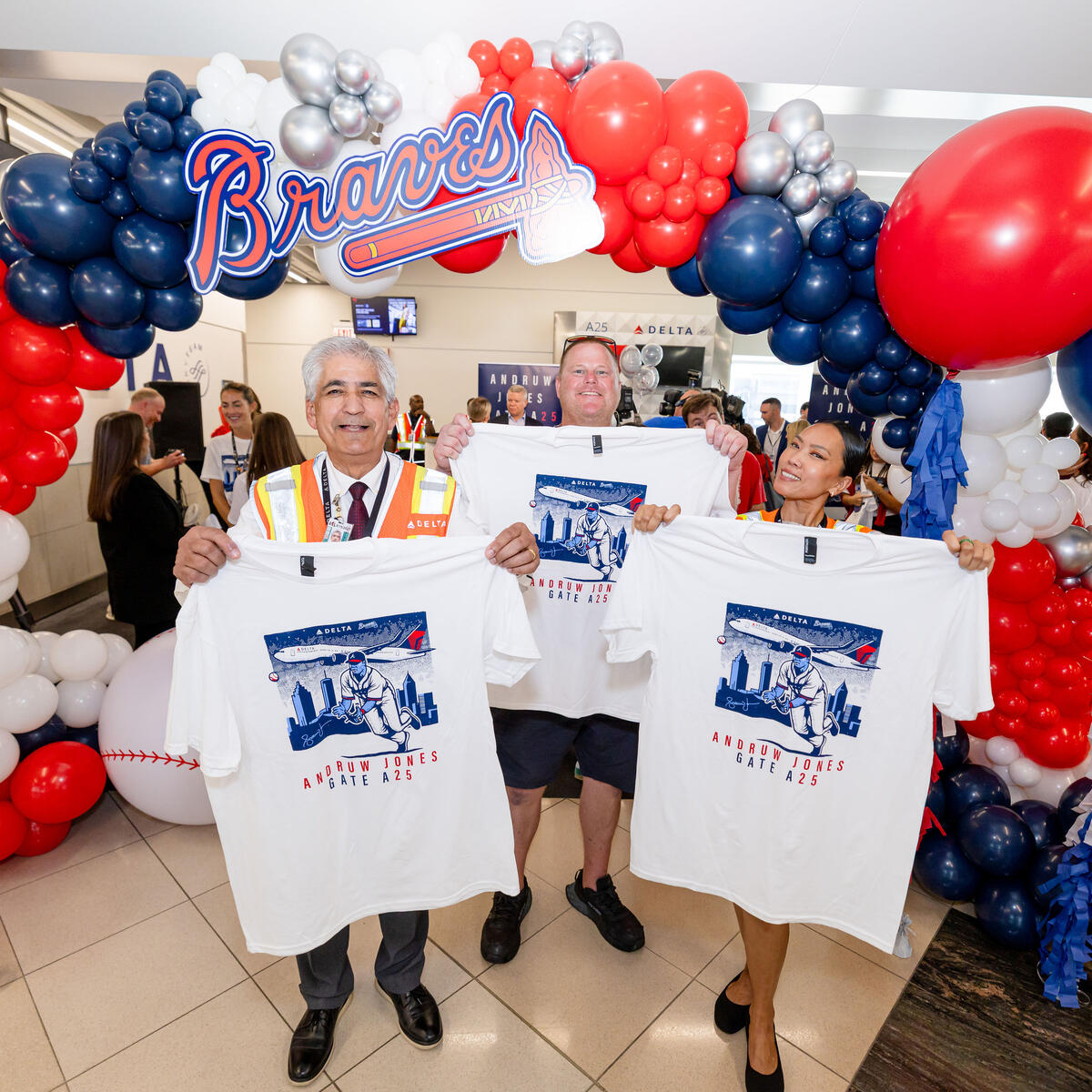 Three attendees stand beneath a Braves-themed balloon arch, displaying commemorative Andruw Jones Gate A25 T-shirts at a Delta airport celebration.