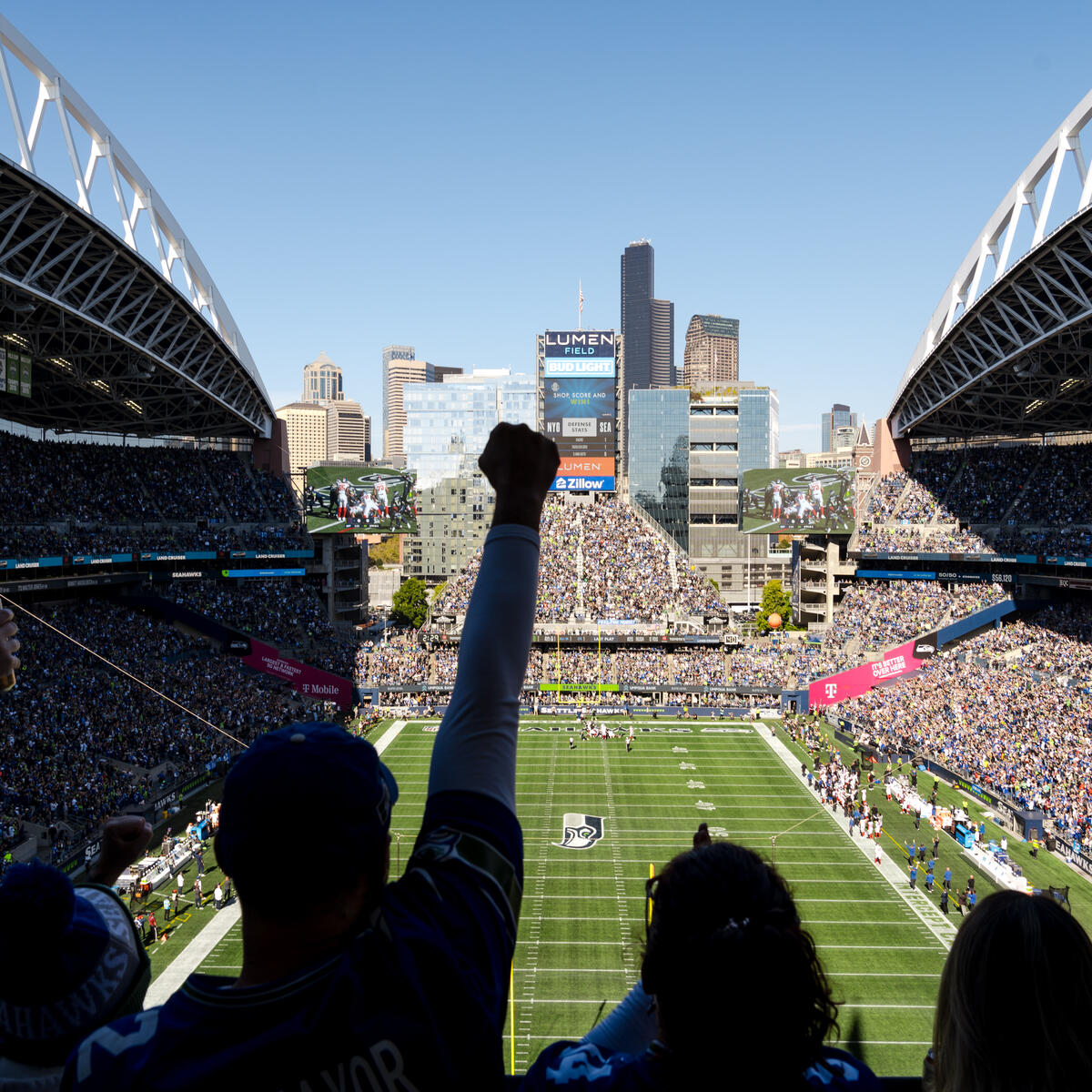 Fans in a packed football stadium watch the game from the stands, with the field and Seattle skyline visible beyond the open end of the venue.