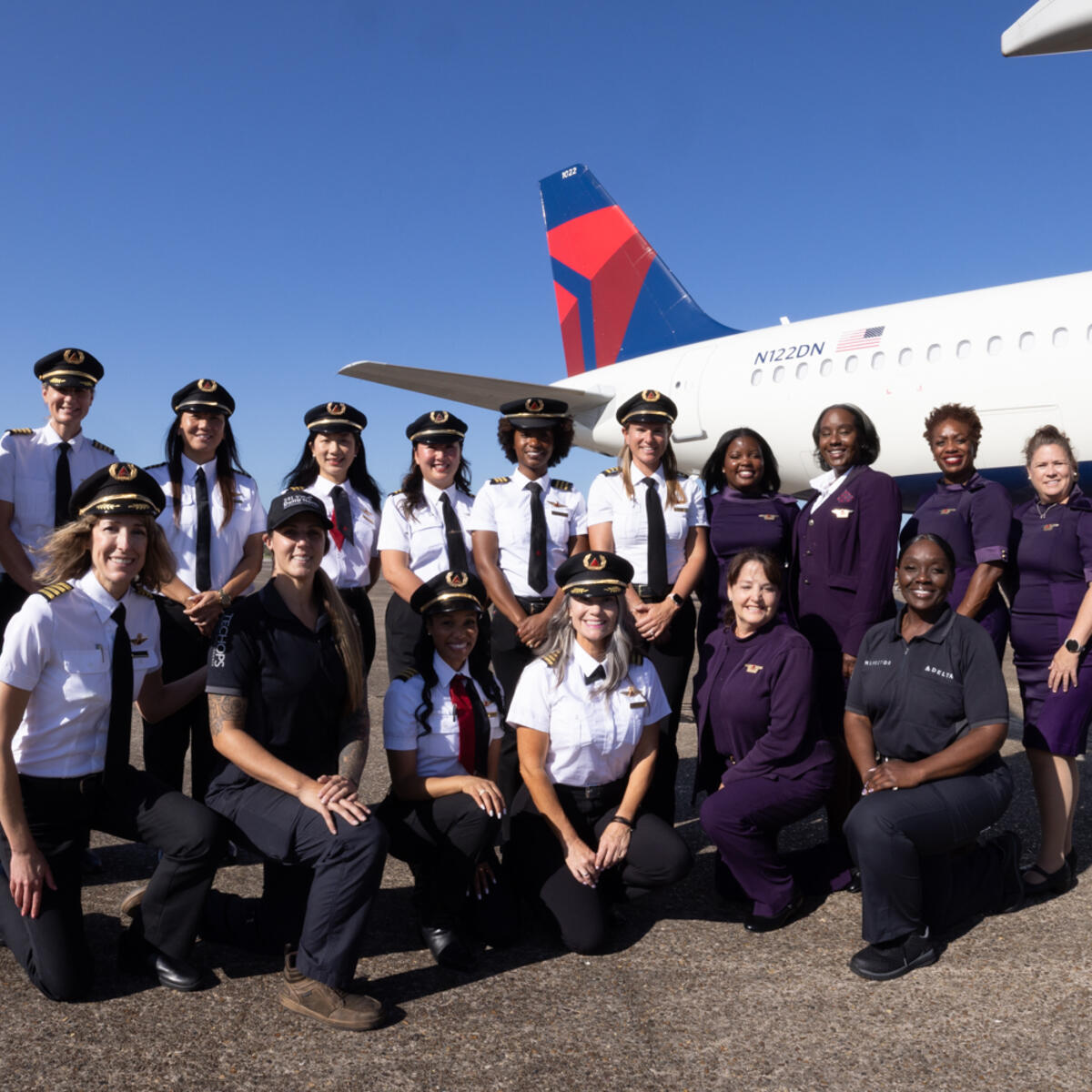 Group of aviation professionals and participants posing on the tarmac in front of a Delta aircraft under a clear blue sky during a Girls in Aviation Day event.