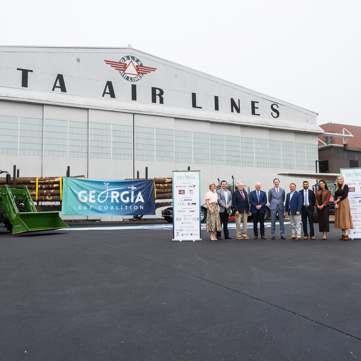 Group stands in front of Delta Air Lines’ historic hangar at the Delta Flight Museum alongside a tractor, timber load and Georgia SAF Coalition signage during a sustainable aviation fuel event.
