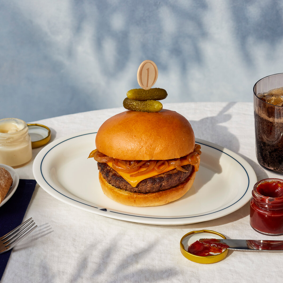 Cheeseburger topped with caramelized onions and pickles, served with condiments, bread, and a soft drink on a white tablecloth.
