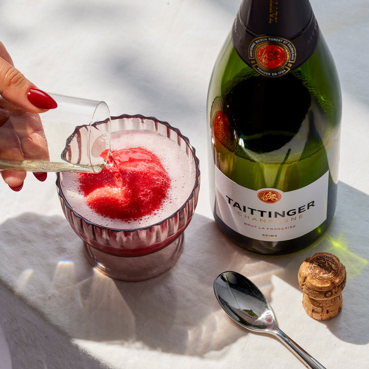 A hand pours sparkling wine over a red fruit sorbet dessert beside a champagne bottle on a white tablecloth.