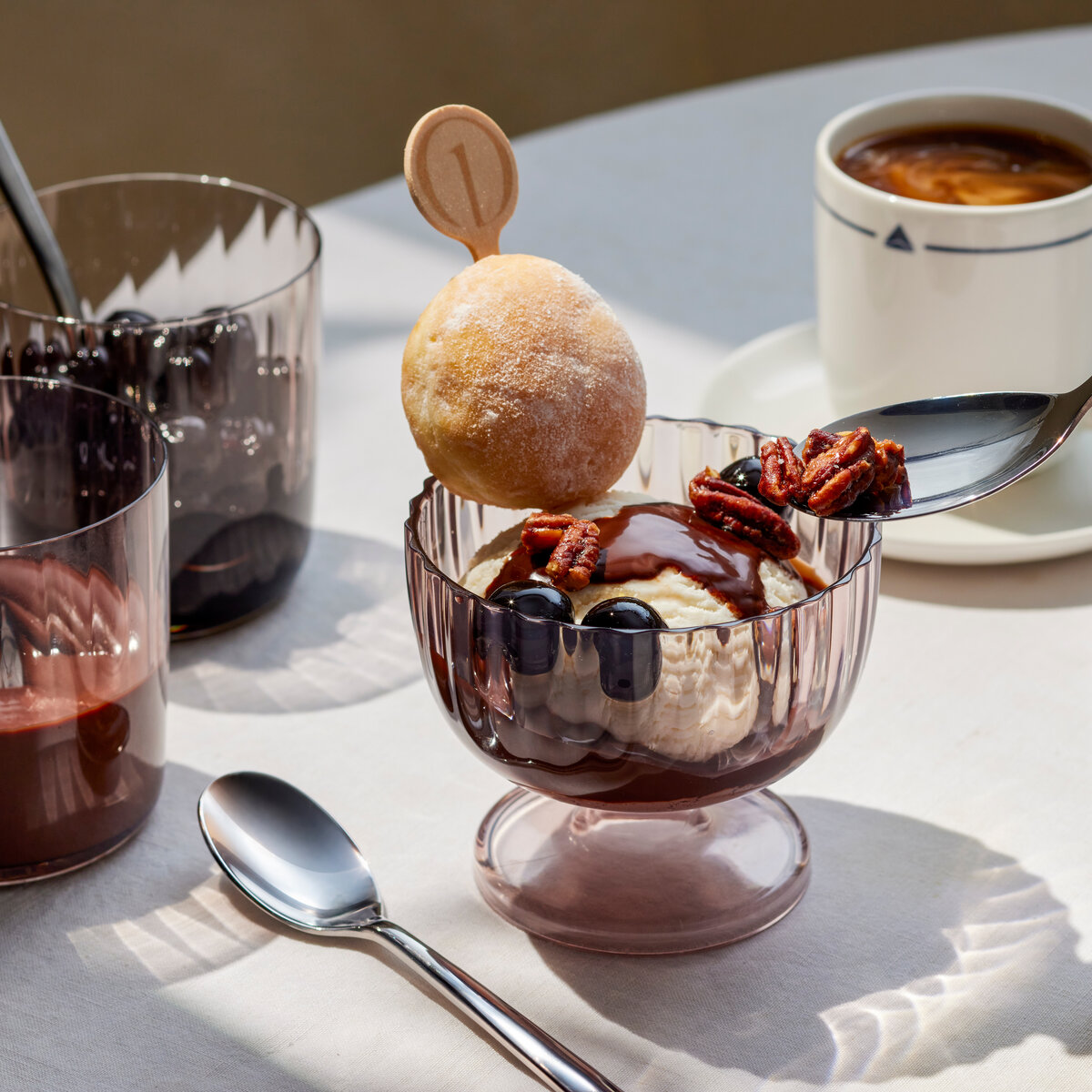 Ice cream sundae topped with chocolate sauce, pecans, and cherries, served with coffee and dessert accompaniments on a table.