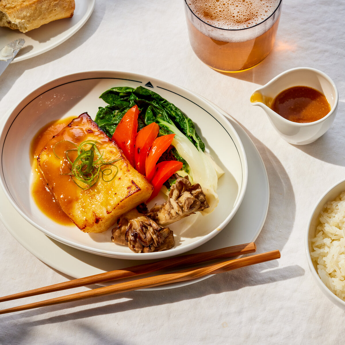 Plated glazed fish with vegetables, rice, bread, and sauce served on a white tablecloth with chopsticks and a beverage.