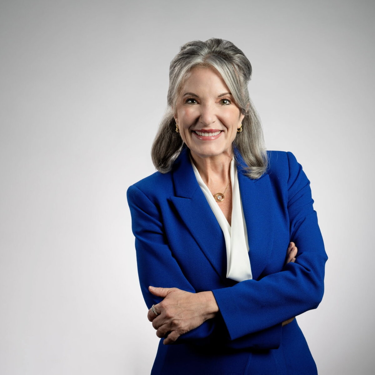 Studio portrait of a person wearing a blue blazer and white blouse, standing with arms crossed against a light gray background.