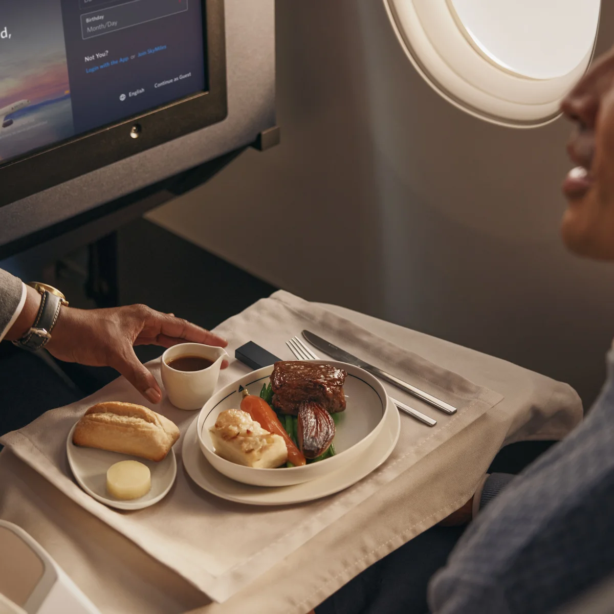 Flight attendant serves a plated meal with bread and vegetables to a passenger in a Delta One cabin.