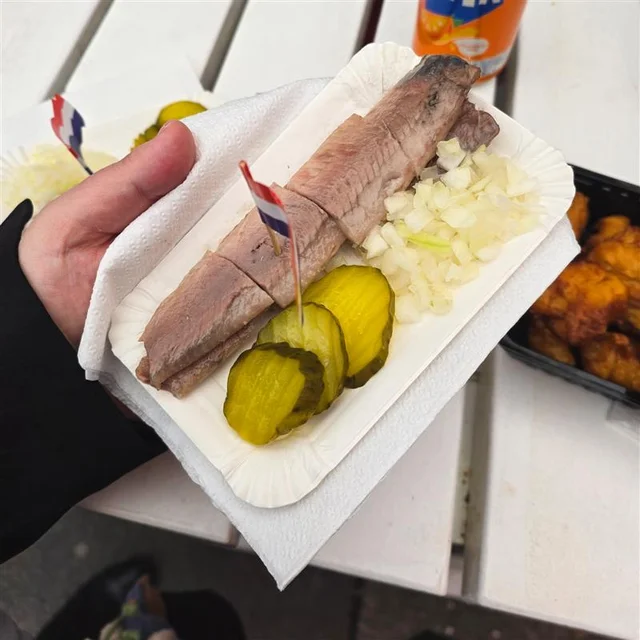 tray of Dutch-style raw herring served with chopped onions and pickle slices, held in one hand with a small Dutch flag toothpick.