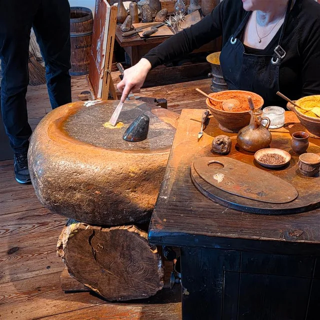 A person uses a stone grinding wheel at a rustic workbench covered with bowls of pigments, tools, and containers.
