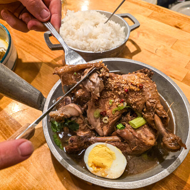 A hand serves braised chicken from a metal pan topped with green onions and spices, alongside a halved boiled egg and a bowl of white rice on a wooden table.