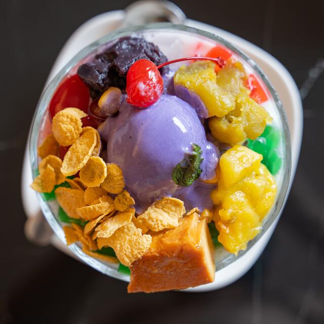 A colorful halo-halo dessert topped with purple ube ice cream, sweet fruits, cornflakes, jelly, and a cherry in a glass bowl.