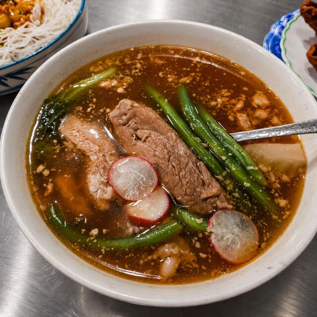 A bowl of savory soup with beef, green beans, radish slices, and leafy vegetables in a rich broth, served with noodles on the side.