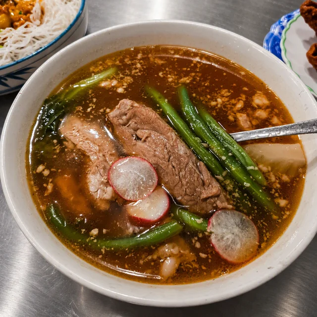 A bowl of savory soup with beef, green beans, radish slices, and leafy vegetables in a rich broth, served with noodles on the side.