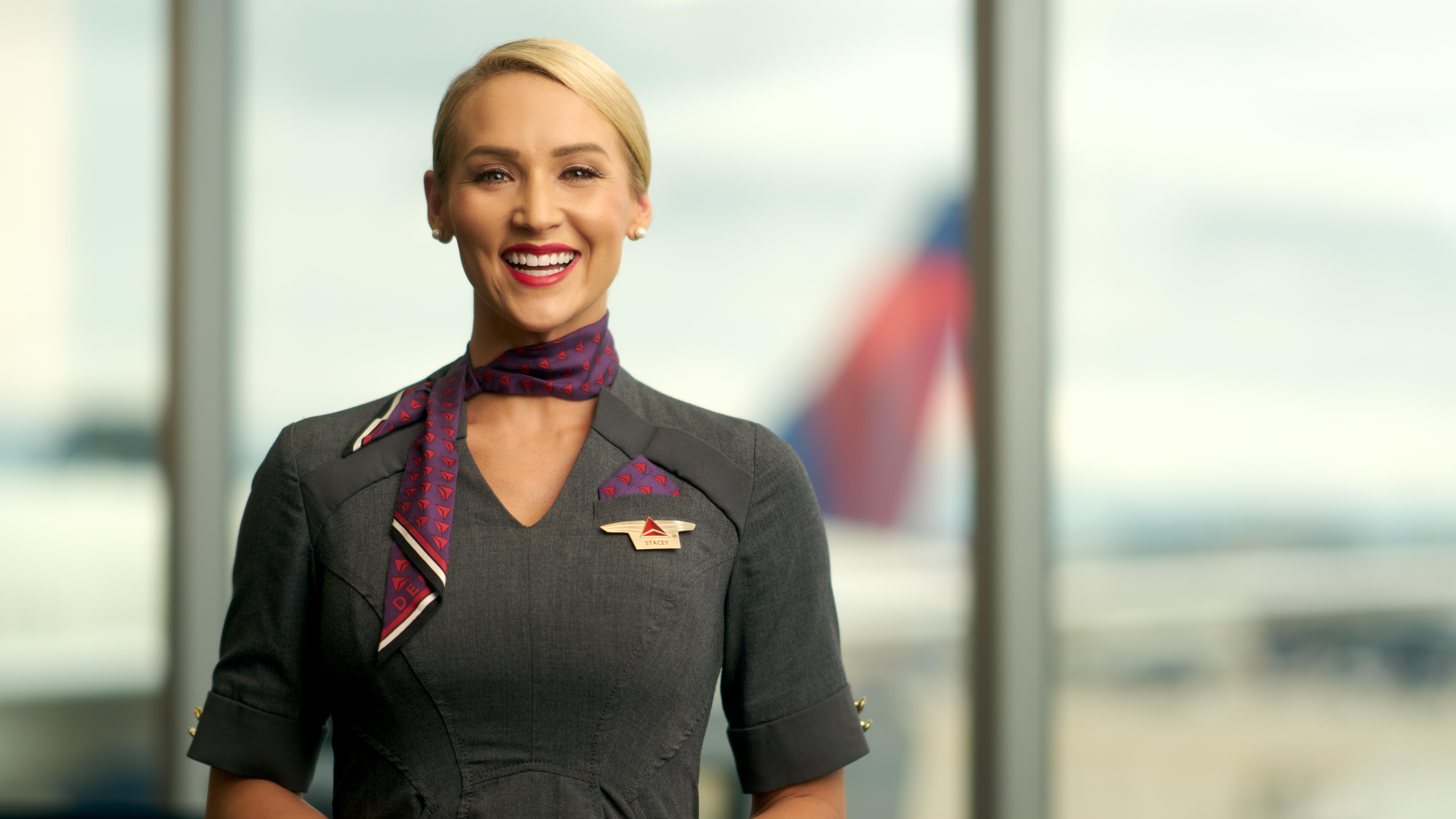 Delta flight attendant smiling, wearing gray dress and purple neck scarf.