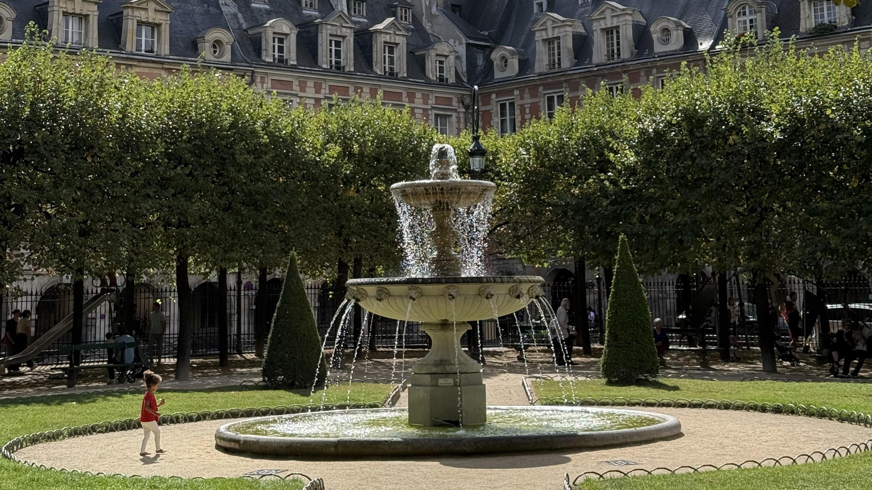Stone fountain in a leafy Paris square, framed by manicured trees and historic red-brick buildings with slate roofs.