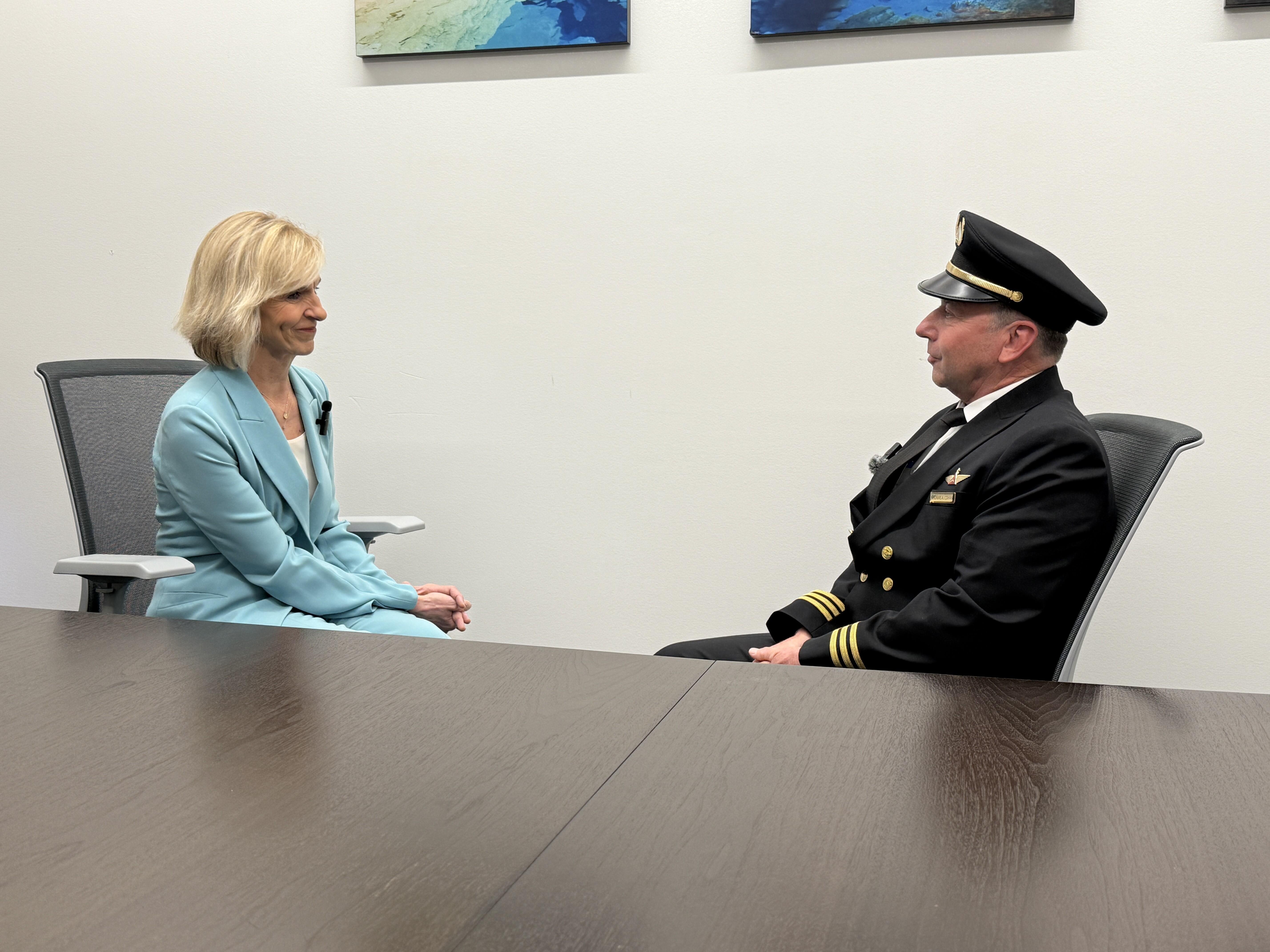 Allison Ausband and Tom Cohan sit facing each other at a conference table, Allison in a business suit and Tom in a pilot uniform.